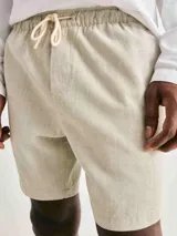 Bermuda de hombre color beige claro, con cintura elástica y cordón de ajuste. El tejido presenta una textura visible, similar al lino o una mezcla de algodón texturizado.