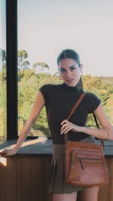 Bandolera marrón de símil cuero con dos bolsillos frontales con cierre, detalles de tachas metálicas, un bolsillo trasero y tres bolsillos internos. Incluye correa larga regulable.