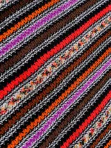 Capa tipo poncho de tejido de punto con diseño de rayas verticales en zigzag, en tonos marrones, negros, naranjas, rojos y morados. Presenta cuello en V y flecos en el ruedo.