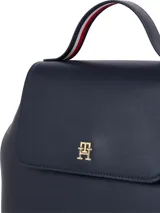 Mochila de poliuretano color azul marino con silueta estructurada y cierre de solapa con broche magnético. Presenta un asa de mano superior y correas de hombro ajustables con cinta distintiva de Tommy Hilfiger. El frente lleva un monograma TH metálico en dorado.