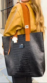 Bolso tote negro con textura símil piel de cocodrilo, doble asa de cuero color marrón y cierre con botón a presión.