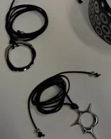 Collar tipo choker de varias tiras de cinta negra de gamuza o terciopelo, anudado al frente con un dije grande de metal plateado con forma de anillo irregular y orgánico.