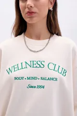 Buzo de algodón color crudo con cuello redondo y manga larga. Presenta bordado frontal en color verde que dice "Wellness Club Body. Mind. Balance Since 1994".
