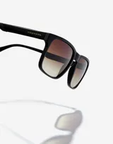 Lentes de sol Hawkers Peak Metal unisex, con montura cuadrada negra y lentes degradadas de marrón a transparente.