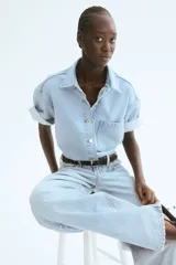 Camisa celeste de denim, corte amplio, con cuello clásico, manga corta, cierre frontal con botones metálicos, bolsillo de parche en el pecho y canesú en la espalda.
