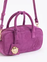 Cartera tipo baulito de cuero descarne vacuno en color fucsia, con diseño compacto y estructura rígida. Presenta doble asa de mano, logo de la marca grabado en relieve en el frente y un charm metálico en forma de vaquita de San Antonio colgando de una de las asas.