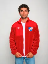 Campera polar roja con cierre frontal, cuello alto y bolsillos laterales. Presenta un panel liso en la parte superior del pecho y un bolsillo con cierre en el lado izquierdo. Incluye el logo de Umbro bordado en el pecho izquierdo y el escudo del Club Nacional de Football bordado en el pecho derecho.