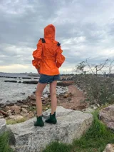 Chaqueta impermeable naranja con cuello alto y detalles de volados en color verde oliva. Presenta cierres y bolsillos frontales, puños ajustados y cordón regulable en la cintura.