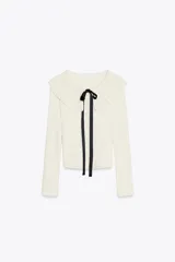 Chaqueta blanca entallada de punto con cuello bobo de tricot y manga larga. Tiene una lazada negra en el cuello y cierre frontal con botones.