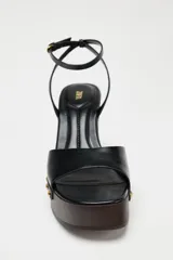 Sandalias negras con plataforma de madera, tacón de cuña y tira delantera con detalle de tachas doradas. Cierre mediante tira con pulsera al tobillo y acabado en punta cuadrada.