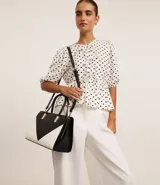 Bolso satchel mediano bicolor, negro en la parte superior y blanco en la inferior, con detalles metálicos dorados en las asas. Incluye correa larga desmontable y cierre de cremallera.