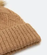 Gorro de lana color camel con textura en punto trenzado y pompón de pelo sintético en la parte superior.