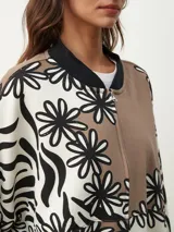 Campera bomber con estampado de flores en blanco y negro sobre fondo beige y marrón. Tiene cuello, puños y bajo acanalados en negro, y cierre frontal con cremallera.