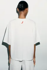Camiseta blanca de algodón con cuello redondo, mangas cortas y hombros caídos. Estampado frontal con el título de la película "El Diablo Viste de Prada" en letras rojas.