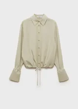 Camisa color beige de corte crop, confeccionada en mezcla de lino y lyocell. Tiene cuello camisero, cierre frontal con botones, manga larga con puños abotonados y lazo ajustable en la cintura.