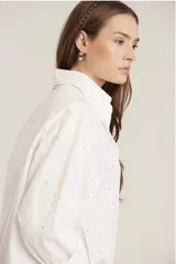 Camisa blanca de corte clásico con cuello abotonado, cierre frontal con botones y mangas largas con puños abotonados. Presenta pequeños apliques de strass plateados dispersos por toda la prenda.