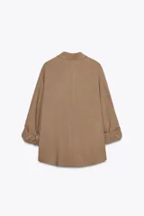 Camisa color marrón claro de cuello solapa y manga larga con trabilla y botón para remangar. Tiene un bolsillo de plastrón en el delantero, bajo asimétrico y cierre frontal con botones ocultos por solapa.