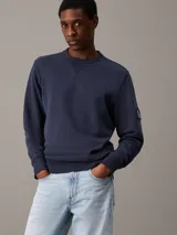 Buzo azul marino de algodón con cuello redondo y ribete en V. Tiene puños y dobladillo acanalados, y una insignia con el monograma de Calvin Klein en la manga.