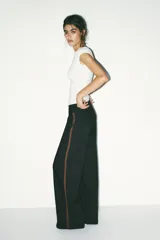 Pantalón negro con finas rayas verticales blancas, de tiro medio y corte ancho. Presenta bolsillos delanteros y traseros, cintura doble tipo bóxer con botones y cierre frontal con cremallera y ganchos metálicos. Detalle de franja marrón en los laterales.