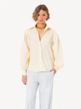 Camisa amarilla confeccionada en poplin rayado con hilos de lurex, mangas abullonadas, botones visibles forrados a tono de tela y detalles de remache Lipstick en la punta del cuello.