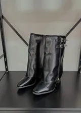 Botas de caña media negras de cuero sintético, con punta cuadrada, taco bajo y cierre lateral con cremallera. Presentan dos correas con hebillas metálicas decorativas en el lateral exterior.