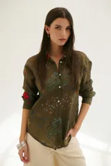 Camisa beige de manga larga con cuello clásico y cierre frontal con botones. Presenta bordados con motivos florales y geométricos en tonos azules, rojos y verdes.
