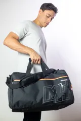 Bolso deportivo tipo duffel bag, color negro, con grandes iniciales 'MH' en el frente en tono gris oscuro. Presenta detalles de franjas horizontales en rojo, amarillo y negro sobre los cierres de los compartimentos. La correa de hombro lleva la inscripción 'NBA' en relieve.