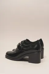 Zapato de cuero vacuno charol negro, con punta redondeada y detalle de perforado en la capellada. Tiene una tira con hebilla metálica y un taco cuadrado de 4 cm.