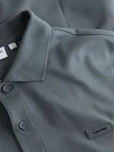 Chomba Calvin Klein gris de corte slim, confeccionada en algodón orgánico. Presenta cuello clásico con solapa de dos botones, mangas cortas con puños de punto, aberturas laterales y logo tonal bordado en el pecho.