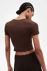 Remera marrón de punto elástico, corte cropped y mangas cortas. Presenta costuras fruncidas en un hombro y a ambos lados de la cintura, creando un efecto drapeado.