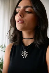 Collar con tiento negro y dije plateado en forma de flor con centro texturizado.