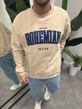Buzo beige de corte holgado con cuello redondo y puños acanalados. Estampa frontal con la palabra "Bohemian" en azul y la frase "Est. 2014 Design" en negro.
