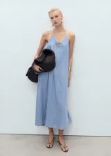Vestido largo de mezclilla, color azul claro, con escote de pico y tirantes finos ajustables. Presenta un detalle de lazo en el centro del escote y un corte recto que se ensancha ligeramente hacia el ruedo.