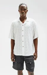 Camisa blanca con bordados calados formando un patrón de hojas y puntos. Tiene cuello clásico, manga corta y cierre frontal con botones marrones.