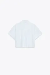 Camisa blanca corta con rayas verticales celestes, cuello solapa y mangas cortas con dobladillo. Tiene un bolsillo de parche en el pecho y cierre frontal con botones ocultos.