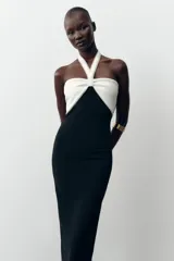 Vestido midi negro de tejido elástico, con escote recto y cuello halter. Presenta un lazo blanco a contraste en el delantero con aplicación de perlas y espalda descubierta.