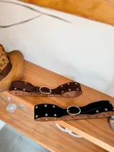 Cinturón ancho de doble capa, con capa superior de gamuza negra o marrón y capa inferior de cuero color camel. Presenta múltiples ojales metálicos y hebilla ovalada metálica de color dorado.