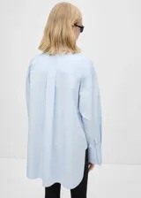 Camisa celeste de algodón, corte oversize, con cuello camisero, mangas largas con puños abotonados y cierre de lazo en el cuello.
