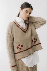Sweater beige con cuello en V y mangas largas con diseño trenzado. Tiene dos bolsillos frontales con bordados de tréboles rojos y ribetes rojos en cuello, puños y ruedo.