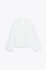 Camisa blanca de cuello redondo y escote pico, con mangas largas abullonadas y detalle de puntillas combinadas a tono en el delantero. Cierre frontal con botones forrados.