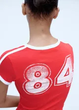 Remera roja de corte recto con cuello redondo y mangas cortas con terminaciones en contraste blanco. Presenta dos líneas blancas paralelas en los hombros.