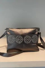 Sobre de cuero vacuno color bronce metalizado, con diseño de cuatro círculos formados por remaches metálicos plateados en el frente. Cuenta con correa larga ajustable, cierre superior y bolsillo trasero con cierre.