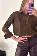 Camisa corta marrón de cuero ovino desgastado con cuello clásico, cierre frontal con botones y mangas largas con aberturas.