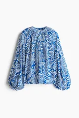 Blusa con estampado de hojas en azul y blanco, de tejido liviano, con cuello corto fruncido y tapeta de botones. Mangas globo largas con volumen de corte raglán y puños finos abotonados.