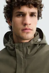 Parka verde militar de tejido técnico ligero resistente al viento, con cuello alto, capucha y manga larga con puños ajustables. Tiene bolsillos de plastrón con solapa en la cadera, bolsillo interior y bajo ajustable con elásticos. El cierre frontal es con cremallera oculta por solapa con botones a presión.
