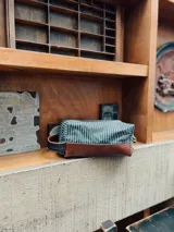 Neceser rectangular con cuerpo superior a rayas verticales blancas y azules, base de cuero marrón y manija lateral de cinta negra.