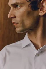 Camisa blanca de algodón con cuello italiano, tapeta lisa, canesú en la espalda y bajo redondeado. Mangas largas con tapeta de manga y botón, y cierre ajustable de botón en los puños. Corte ajustado que realza el contorno del cuerpo creando una silueta entallada.