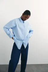 Camisa celeste de algodón suave, de corte holgado, con cuello clásico, botones frontales, canesú en la espalda, hombros caídos, bolsillo de parche en el pecho, mangas largas con puños abotonados y bajo redondeado.