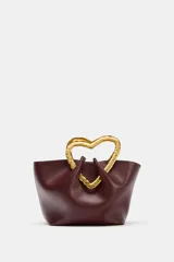 Bolso de mano tipo saco, color borgoña, con asa superior metálica con forma de corazón martillado en tono dorado. Incluye un compartimento interior extraíble y cierre mediante botón metálico.