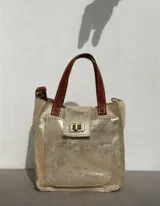 Mini bolso de mano color dorado metalizado con efecto desgastado. Posee dos asas cortas de cuero marrón oscuro con costuras visibles y un cierre de broche metálico en la solapa frontal. Incluye una correa adicional para llevar al hombro.